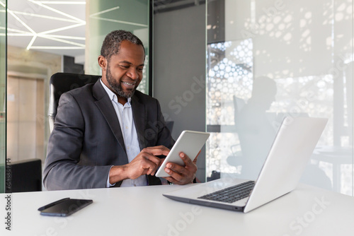 Mature adult african american boss investor at workplace using tablet computer, man happy with financial results and achievement smiling, using app inside office at table.