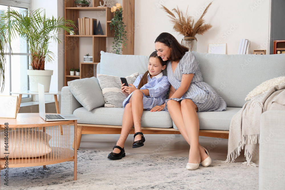 Fototapeta premium Mother and daughter with mobile phone resting on grey sofa in living room