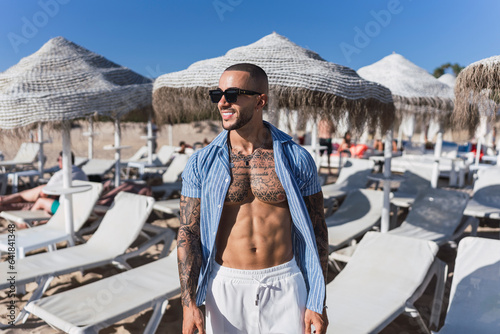 Bild auf Leinwand Chico joven tatuado y musculoso posando en verano en playa soleada