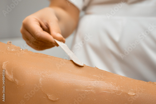 Close up beautician hand applying the epilation gel with a spatula on the female leg. Laser hair removal procedure. 