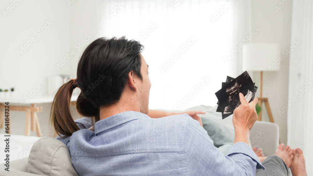 Asian dad and mom couple family looking at pregnant ultrasound of baby image and proud of new born after checkup health care while sitting on sofa in the weekend at house