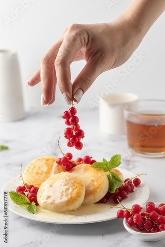 Cottage cheese pancakes or syrniki with red currant and milk sauce on white plate marble background. Woman hand is decorating the syrniki wth berries
