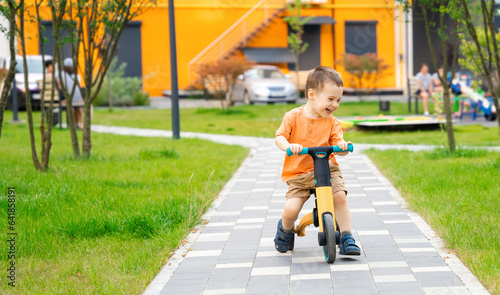 A cute happy toddler boy of two or three years old rides a bicycle or balance bike in a residential complex on a sunny summer day. Active kid playing outside.