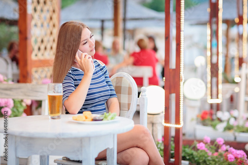 Portrait of a young woman drinking beer on the terrace of a beach cafe and using smartphone