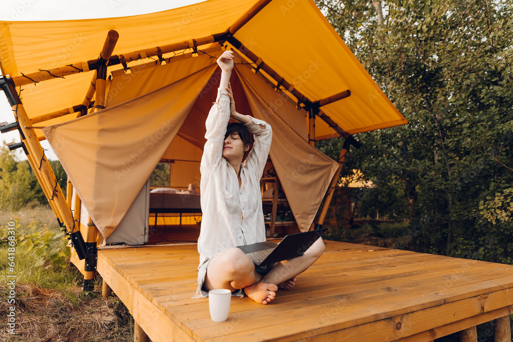 Beautiful woman in cozy outfit works at laptop while sitting on chair near the tent glamping