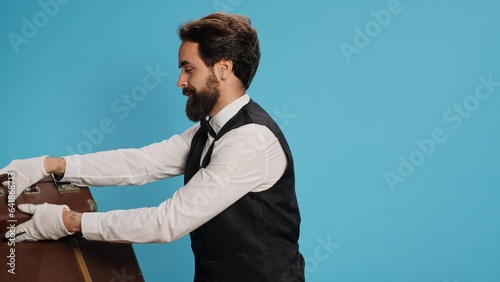 Friendly bellboy indicates satisfaction, showcasing role as professional hotel concierge employee in hospitality industry. Skilled doorman with white gloves gloves carrying suitcase in studio. photo