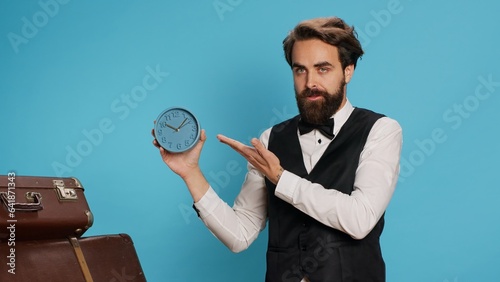 Doorkeeper shows wall clock on camera, checking time and trying to be punctual in hotel travel sector. Man hotel concierge using watch to see hour and minutes in studio next to trolley bags.