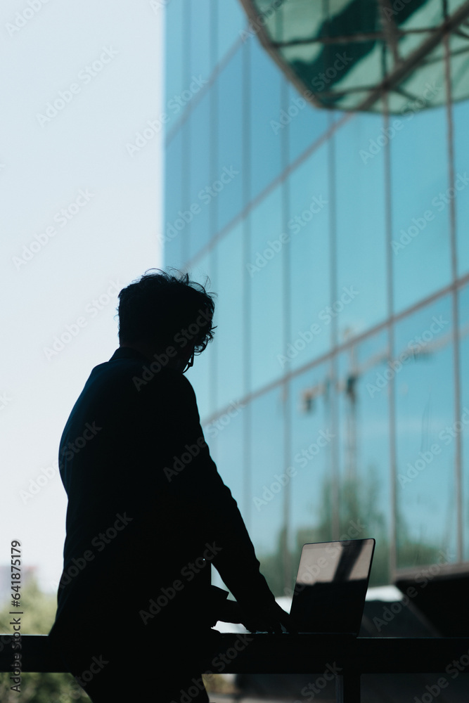 Man using lap top placed on a metal fence in front of big business center. Silhouette photo