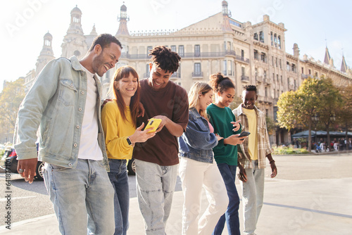 Group of smiling multiracial young people using cell phones. Cheerful students strolling around looking at technological devices. Happy fellow university students on study trip in European city. 