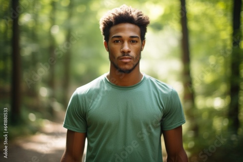 A young African American male standing still wearing a green and white cross country running tshirt and shorts with a defocused background of a winding trail trees and distant