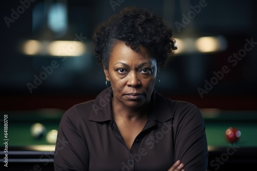 A middleaged African American female billiard player standing still her expression clearly visible against a soft background of billiard cues and table.