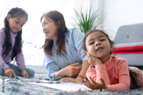 Mother or grandmother relaxing playing with two little girls, lie on fur floor in living room drawing pictures together, one of them smiling at camera