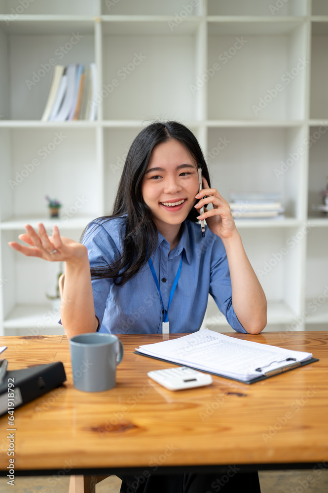 A happy Asian businesswoman enjoys talking on the phone with her friend at her desk A happy Asian businesswoman enjoys talking on the phone with her friend at her desk