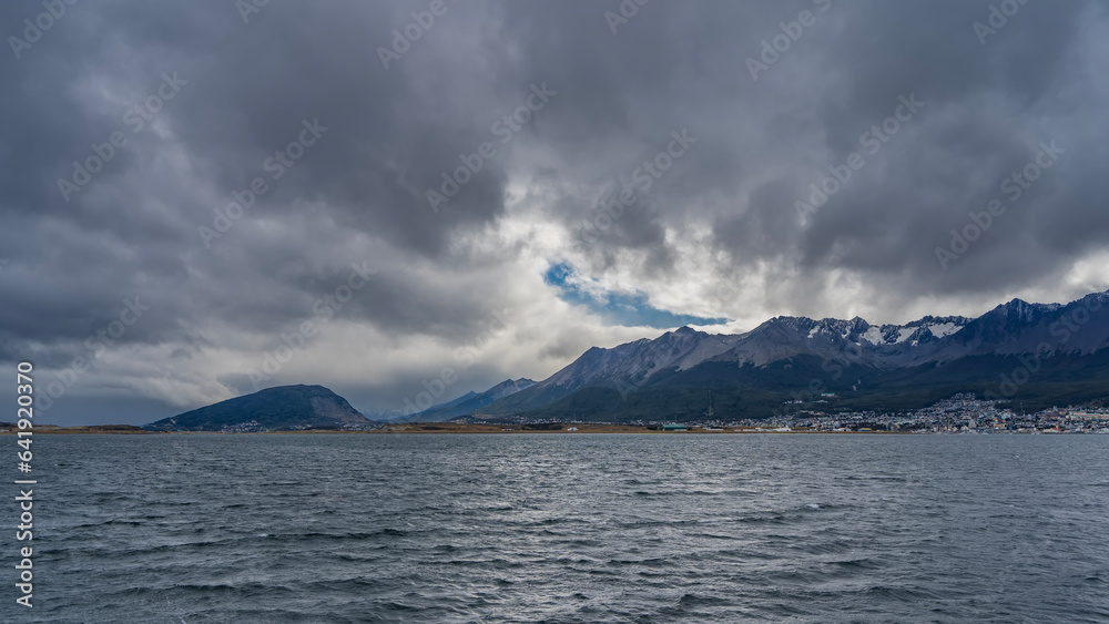 A beautiful mountain range of the Andes against a cloudy sky is visible from the side of the Beagle Channel. City houses of Ushuaia on the shore. Ripples on the blue water. Argentina.