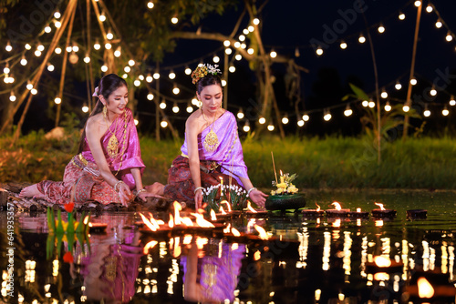 Two thai woman holding a krathong sitting on a raft by the river, Asian women in traditional Thai costumes bring krathongs to float on Loi Krathong Day, traditions and culture of Thailand, © SHUTTER DIN