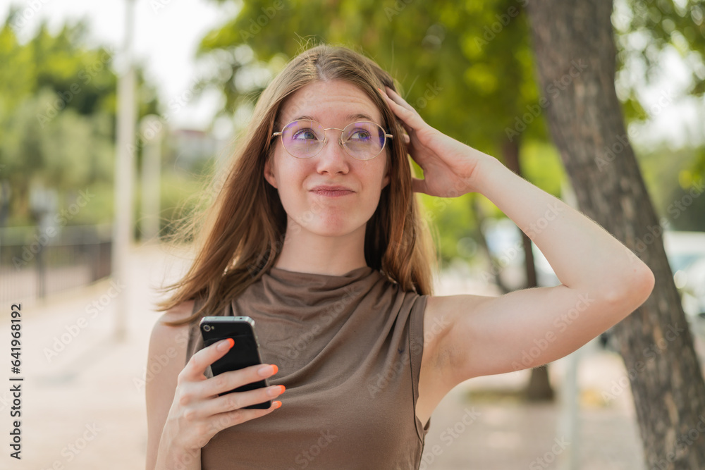 French girl with glasses using mobile phone at outdoors having doubts and with confuse face expression French girl with glasses using mobile phone at outdoors having doubts and with confuse face expression