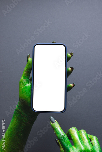 Vertical image of green monster hand holding smartphone with copy space on grey background