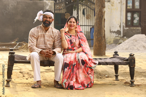 Happy Indian Rural family in village. Husband and wife sitting on cot outside their home front yard. man in kurta pajamas and wife in beautiful saree.