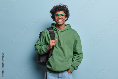 People positive emotions concept. Studio waist up of young happy smiling Hindu male student standing in centre isolated on blue background wearing green hoodie and jeans with black bag on shoulder © Wayhome Studio