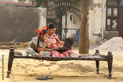 Happy Indian Rural family in village. Rural couple sitting together smiling on cot outside their home in front yard using laptop for online payments.