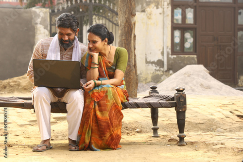 Fotografija Happy rural Indian husband and wife sitting together outside the cottage / home/ house holding a laptop in hands and surfing online