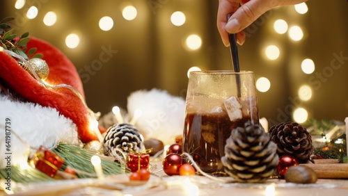 table with Christmas decorations and sweets, stirring hot chocolate with marshmallows. The garland bokeh is behind.