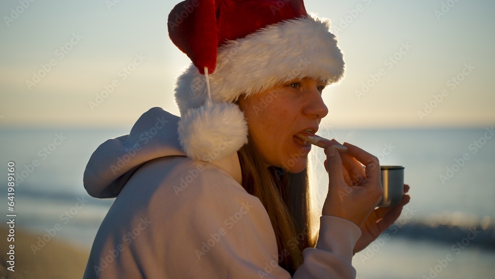 Obraz premium A young woman in a Santa hat celebrates Christmas by the sea with hot tea and a gingerbread cane.