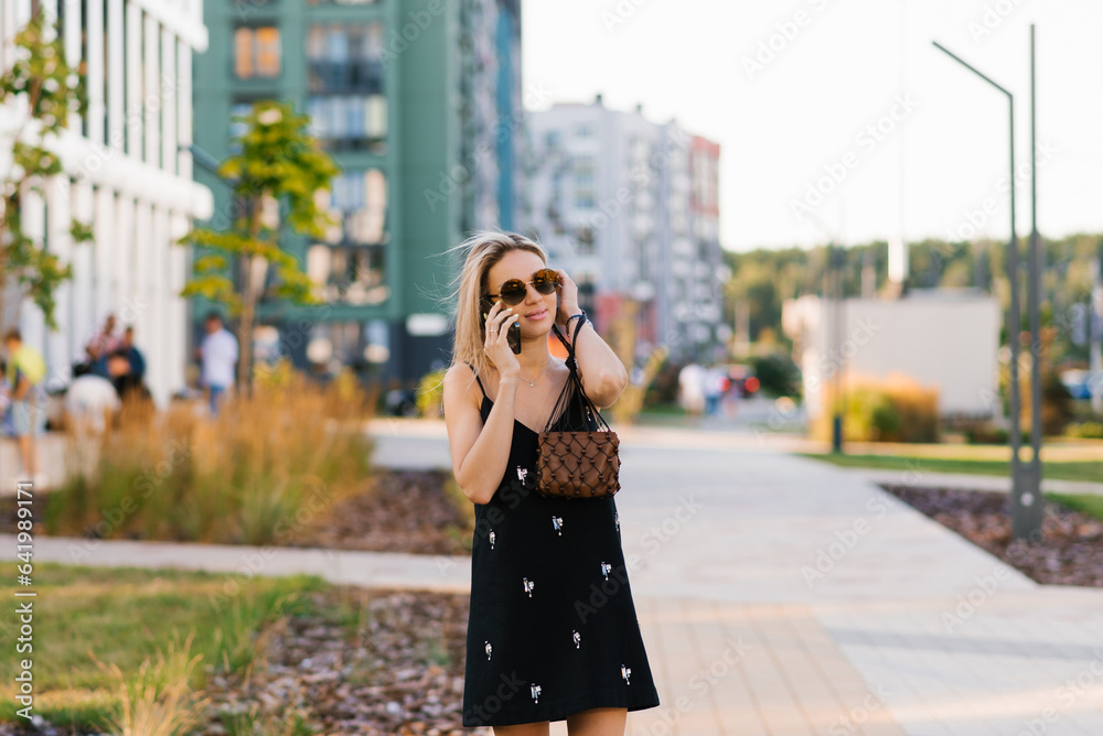 Fototapeta premium Pretty young blonde of Caucasian nationality in sunglasses talks on a smartphone while walking around the city