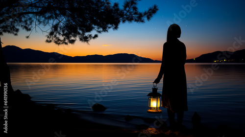 Silhouette of a woman looking into the last light of the day by a lake in the wild, holding a lit vintage kerosene lamp or lantern. Shallow field of view.	