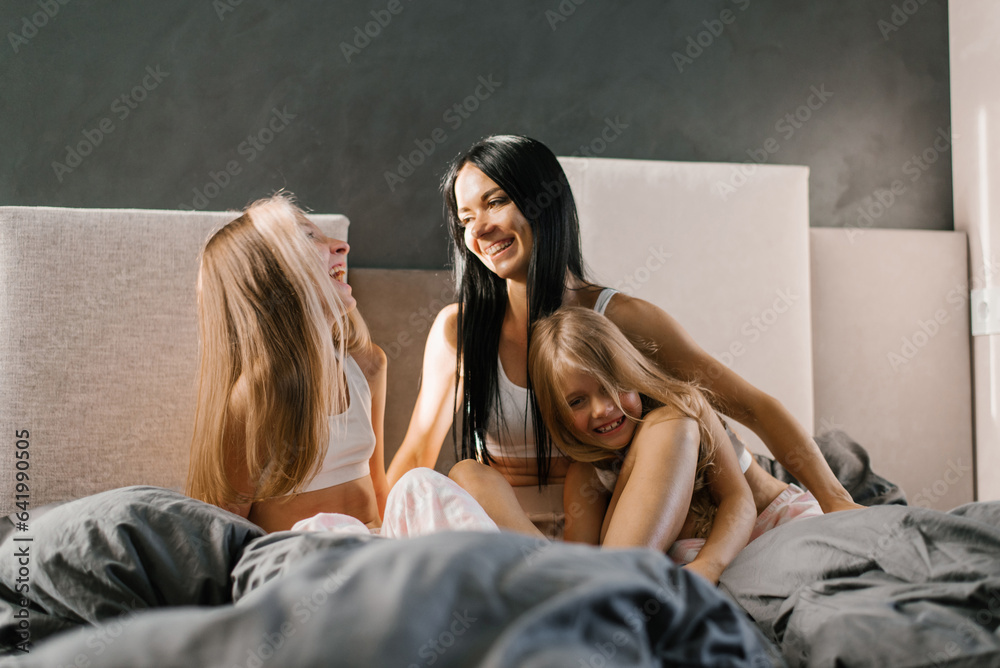 Happy family mom and daughters in pajamas in the morning in bed having fun and chatting - obrazy, fototapety, plakaty 