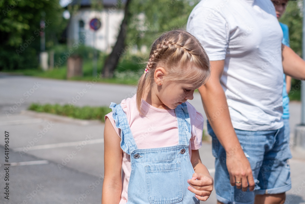A girl with braids stands shyly and interestedly next to her dad and looking down, a parking in the background.