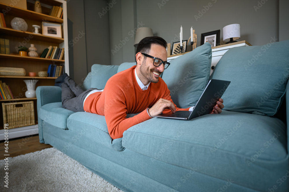 Happy young businessman in glasses reading e-mails while lying comfortably on sofa in living room