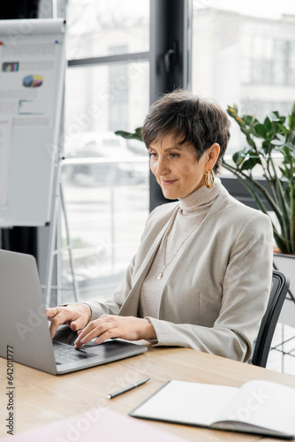 experienced middle aged businesswoman typing on laptop near notebook at workplace in office