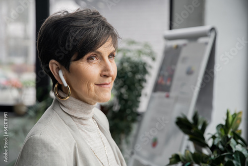 positive and dreamy mature businesswoman listening music in earbud and looking away in office