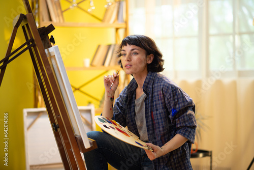 A beautiful woman with a funky pixie haircut is painting something on a large canvas and wearing a blue flannel top that has paint marks on it while being in an art studio