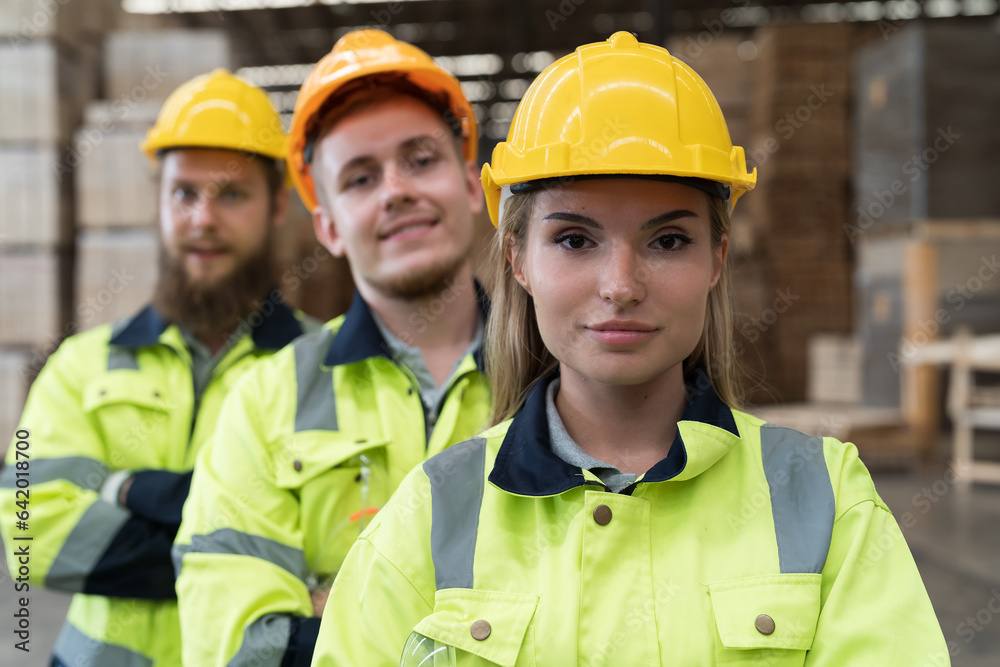 Group of male and female warehouse workers standing together with crossed arms and smiling in industry factory, wearing safety uniform and helmet. Warehouse workers working in warehouse storage