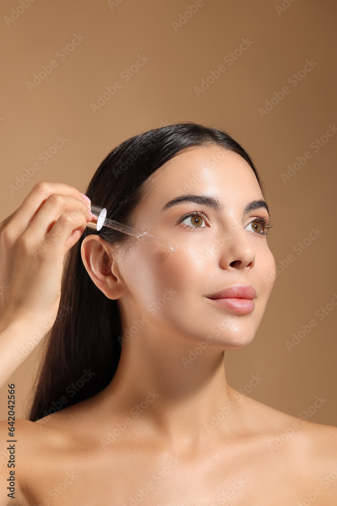 Beautiful young woman applying serum onto her face on beige background