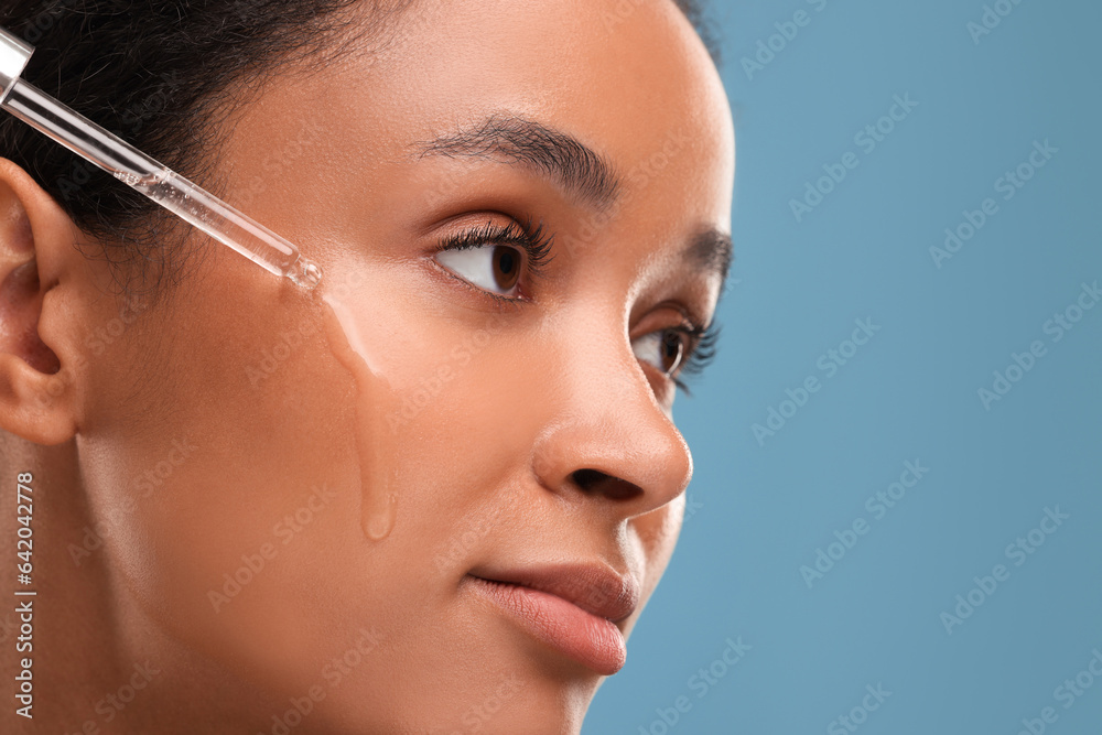 Beautiful woman applying serum onto her face on blue background, closeup. Space for text