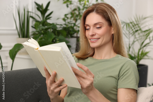 Woman reading book on sofa surrounded by beautiful potted houseplants at home