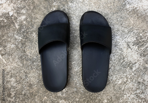 A pair of old black slip-on slippers sandals on grey concrete floor. Basic pair of open toe slide sandals for man, woman, children. Beach slippers for summer vacation.