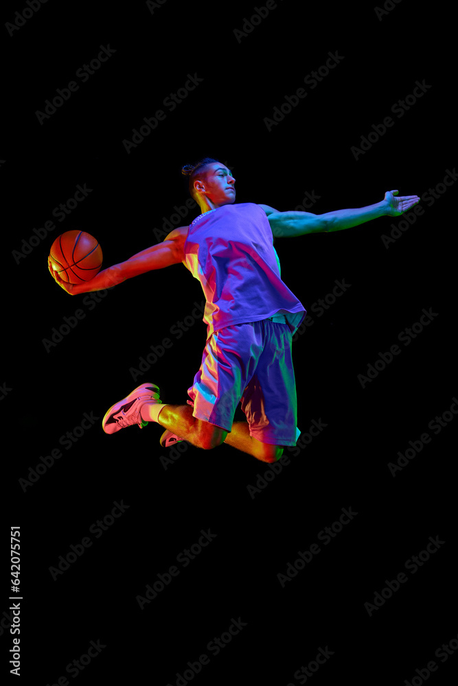 Dynamic image of young man, basketball player in uniform in motion, jumping with ball over black studio background in neon light. Concept of professional sport, competition, hobby, game, competition