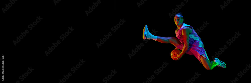 Obraz premium Dynamic image of teen boy during basketball game in motion, jumping with ball against black background in neon light. Concept of professional sport, competition, competition. Banner. Copy space for ad
