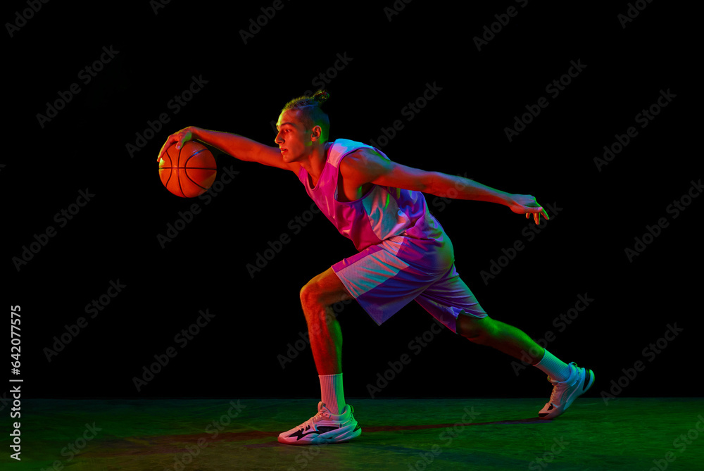 Muscular, active young man in motion, basketball player with ball during game against black background in neon light. Concept of professional sport, competition, hobby, active lifestyle, competition