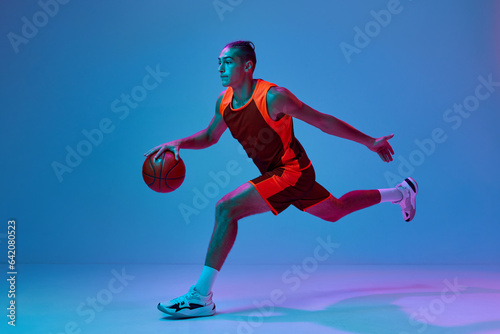 Dynamic image of teen boy in uniform, playing basketball against blue studio background in neon light. Dribbling ball. Concept of professional sport, competition, hobby, game, competition