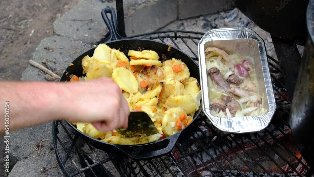 Stirring almost tender potatoes slices and carrots while being cook in cast iron pan over fire pit alongside duck meat odds like neck and gizzard in the aluminum tray.