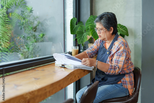A retired Asian senior woman sits in front of a pile of documents. Checking two income and expenditure accounting documents. Use your pension to run a small business, invest with daughter.