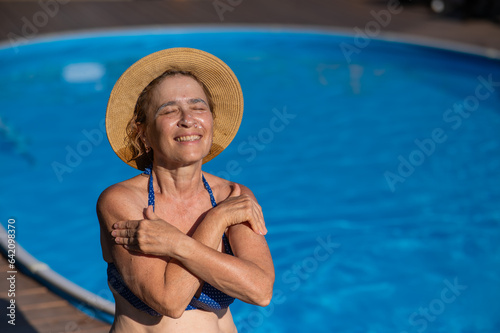An elderly woman in a straw hat is sunbathing by the pool. Retiree on vacation.