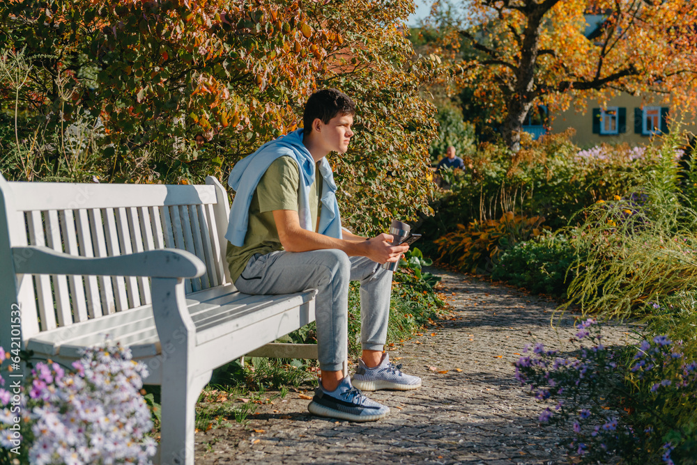A Teenager Sits On A Bench In The Autumn Park Drinks Coffee From A Thermo Mug And Looks Into A Phone. Portrait Of Handsome Cheerful Guy Sitting On Bench Fresh Air Using Device Browsing Media Smm A Teenager Sits On A Bench In The Autumn Park Drinks Coffee From A Thermo Mug And Looks Into A Phone. Portrait Of Handsome Cheerful Guy Sitting On Bench Fresh Air Using Device Browsing Media Smm