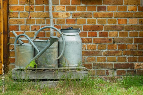Two watering cans and a milk can standing against a brick wall