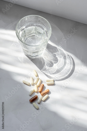 Glass with clean pure water and pills and capsule on white marble table background with aesthetic sunlignt shadow. Healthy morning routine, beauty supplement treatment concept photo